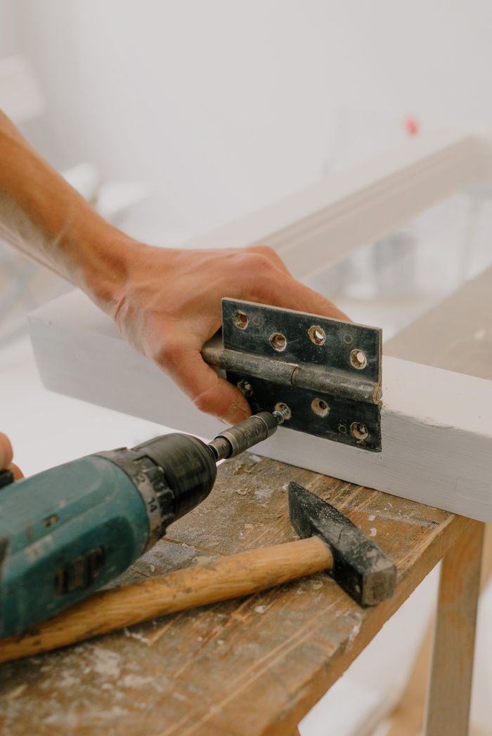 about-04 Crop unrecognizable craftsman attaching metal hinge with modern screwdriver on wooden window frame located on wooden board during renovation works